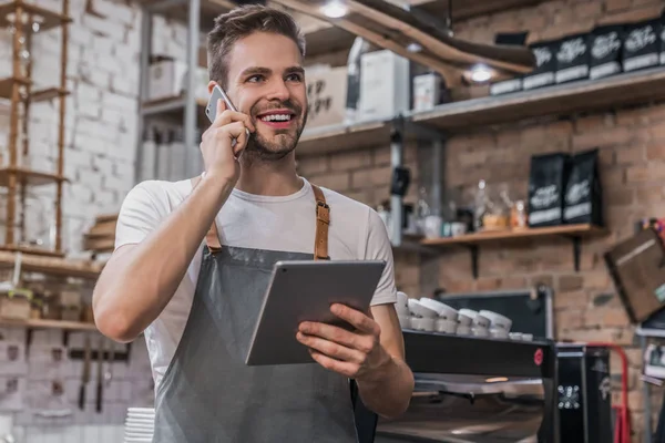 Lachende ondernemer in schort in de buurt van de toonbank van zijn cafe met behulp van digitale tablet en praten op de smartphone — Stockfoto