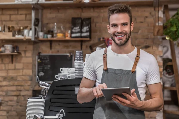 Glimlachende jonge ondernemer draagt een schort in de buurt van de balie van zijn cafe en met behulp van digitale tablet — Stockfoto