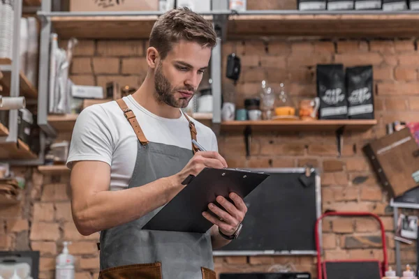 Young male owner writing on clipboard while checking products at coffee shop — Stock Photo, Image