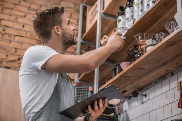 Alacsony látószögű kilátás a fiatal férfi tulajdonos írás vágólapra, miközben kiszámítja termékek kávézóban — Stock Fotó