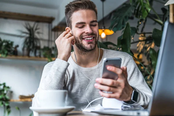 Potret pria yang tersenyum dengan earphone duduk di kedai kopi dan menggunakan ponsel — Stok Foto