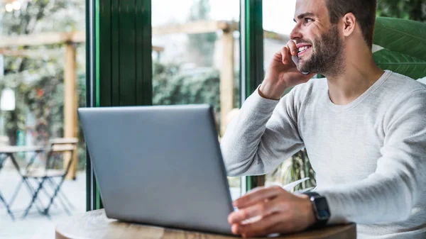 Anak muda yang bekerja di kedai kopi menggunakan ponsel dan laptop saat melihat ke jendela — Stok Foto