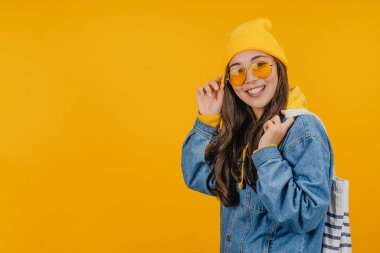 Koyu renk uzun saçlı, sarı şapkalı ve güneş gözlüklü genç bir kadın sarı arka planda tek başına duran bir çanta taşıyor.