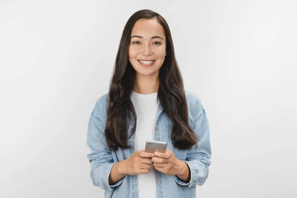 Porträtt av glad flicka i casual håller mobiltelefon och tittar i kameran isolerad över vit bakgrund — Stockfoto