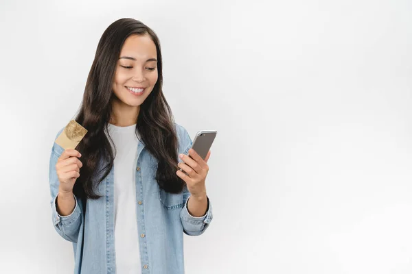 Retrato de niña sonriente sosteniendo el teléfono móvil y mostrando la tarjeta de crédito de plástico de pie sobre fondo blanco — Foto de Stock