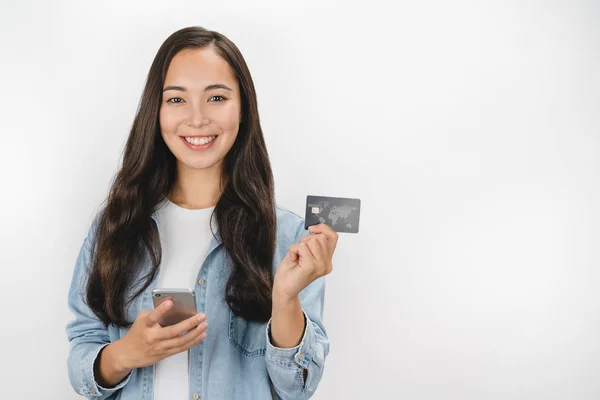 Portrait of smiling asian woman holding credit card and mobile phone while looking at camera isolated over white background — Stock Photo, Image