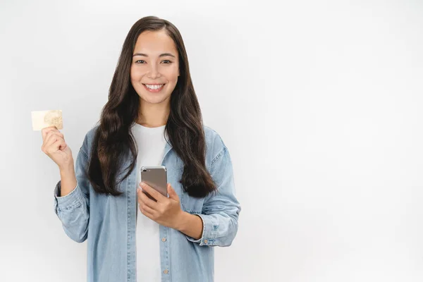 Happy asijské dívka drží mobilní telefon a ukazuje plastové kreditní karty stojící nad bílým pozadím — Stock fotografie