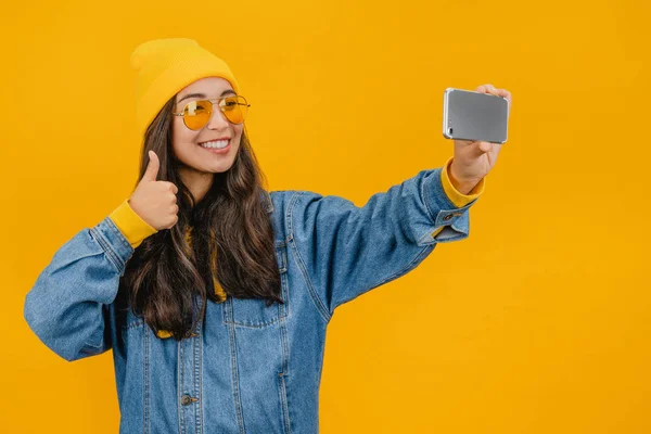 Joven mujer asiática mostrando el pulgar hacia arriba y tomando foto selfie en el teléfono inteligente aislado sobre fondo amarillo — Foto de Stock