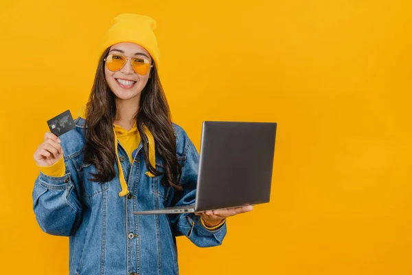 Счастливая женщина в шляпе держит кредитную карту и ноутбук изолированы на желтом фоне — стоковое фото