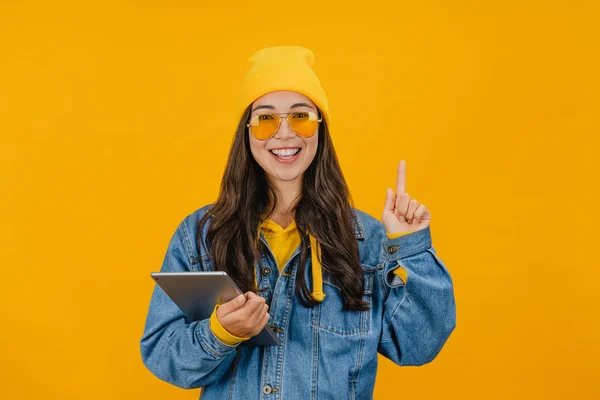 Возбужденная азиатка, держащая в руках цифровой планшет и указывая пальцем на пространство для копирования, стоящая изолированно на желтом фоне — стоковое фото