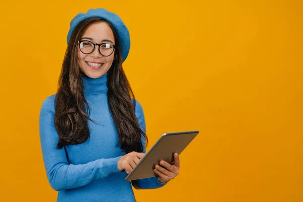 Gadis asia muda berbaret biru menggunakan tablet yang diisolasi dengan latar belakang kuning — Stok Foto