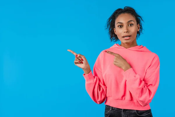 Glückliche junge Afrikanerin zeigt mit dem Finger auf Kopierraum isoliert über blauem Hintergrund — Stockfoto