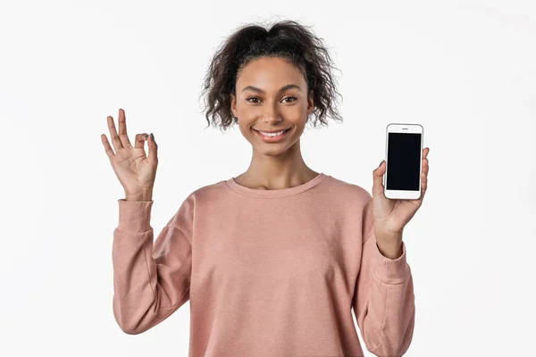 Mujer africana sosteniendo el teléfono inteligente y mostrando signo de ok mientras mira a la cámara sobre fondo blanco — Foto de Stock