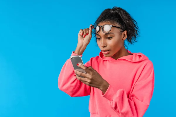 Mavi arka planda cep telefonu tutan gözlüklü Afrikalı genç bir kadın. — Stok fotoğraf