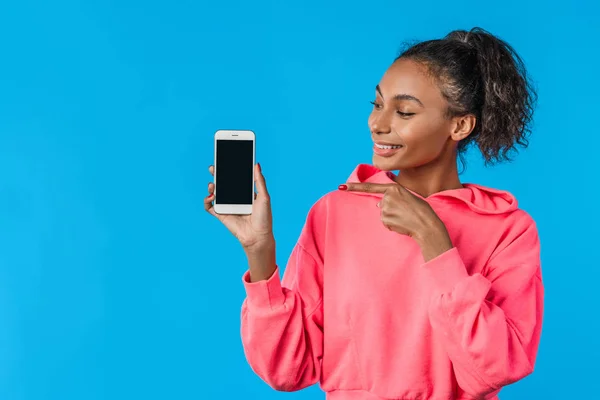 Alegre joven afro mujer sosteniendo el teléfono móvil y señalando el dedo sobre el fondo azul — Foto de Stock