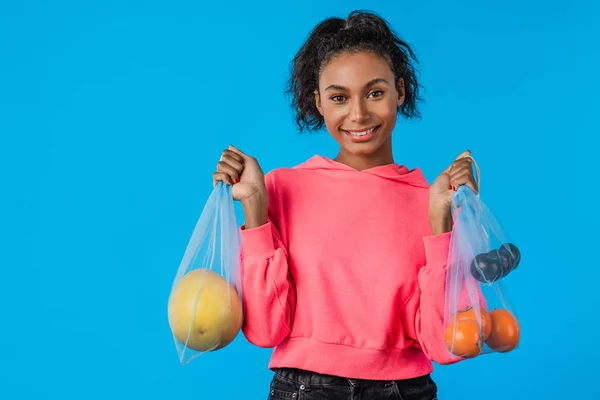 Portret van vrolijk Afrikaans Amerikaans meisje glimlachend en het dragen van nettas met fruit geïsoleerd over blauwe achtergrond — Stockfoto