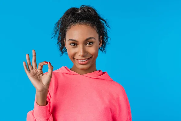 Close up of american woman smiling on camera and gesturing ok sign isolated over blue background — 스톡 사진