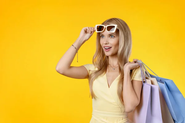Megdöbbent fiatal nő nyári ruha visel napszemüveg pózol bevásárló táskák és félrenéz sárga háttér. — Stock Fotó