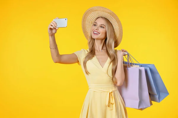 Csodálatos fiatal nő pózol elszigetelt sárga háttér gazdaság bevásárló táskák szelfi mobiltelefonnal. — Stock Fotó