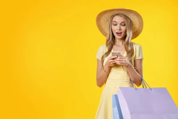 Fénykép a sokkolt nő ruha nézi okostelefon kéz a kézben meglepetés, miközben gazdaság bevásárlótáskák elszigetelt sárga háttér — Stock Fotó