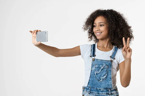Alegre niña africana joven usando el teléfono móvil tomar selfie mostrando gesto de paz posando aislado sobre fondo blanco — Foto de Stock
