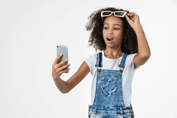 Beyaz arka planda izole edilmiş güneş gözlüklü, cep telefonu kullanan Afrikalı küçük bir kız. — Stok fotoğraf