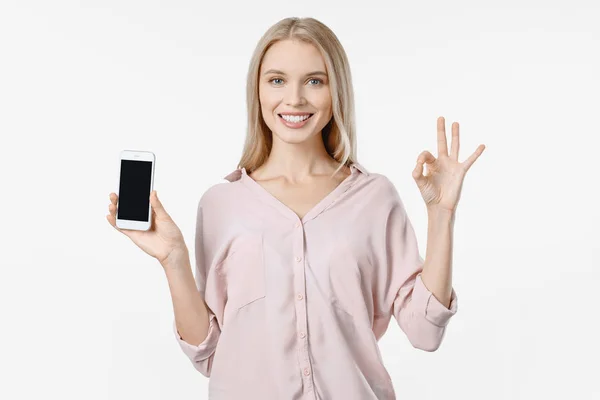 Leende flicka i casual hålla mobiltelefon och visar ok isolerad över vit bakgrund — Stockfoto