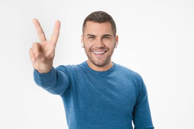 Young man listening to music using earphones smiling with happy face doing victory sign standing over isolated white background