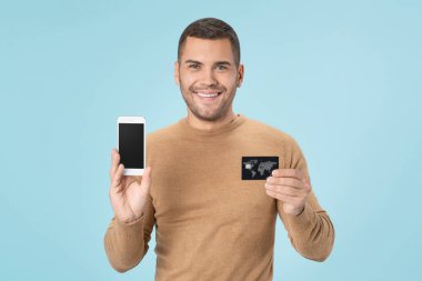 Smiling caucasian man holding credit card and smartphone isolated over blue background