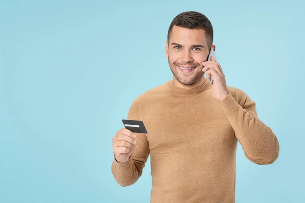 Young smiling man talking on smartphone with credit card in hand on background of blue color — Stock Photo, Image