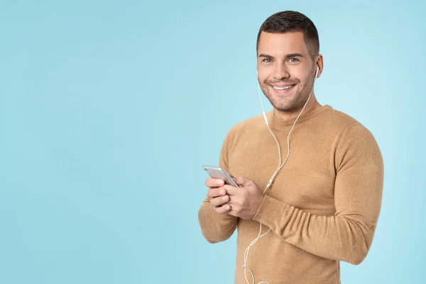 Happy man using mobile phone listening music with earphones posing isolated over blue background — Stok fotoğraf