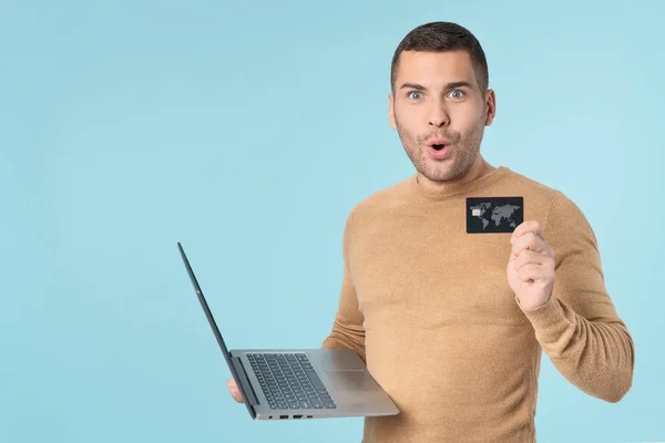 Portrait of surprised handsome man wearing casual clothes using laptop computer and showing credit card standing isolated over blue background — 스톡 사진