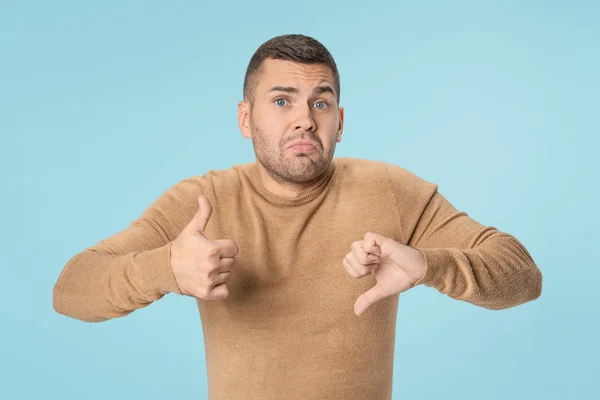 Young man confused with thumbs up and down trying to take a decision over blue background — 스톡 사진