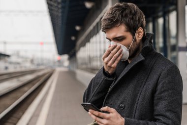 Solunum cihazındaki yakışıklı adam istasyonda treni beklerken akıllı telefon kullanıyor. Enfeksiyon ve hava kirliliğini önleme kavramı