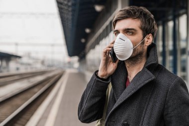 Korumacı maskeli genç beyaz erkek tren istasyonunda cep telefonuyla konuşuyor.
