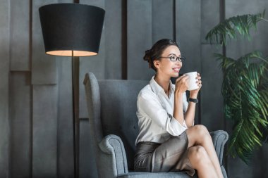 Bir fincan sıcak içecekle düşünceli genç bayan lobi sandalyesinde oturmuş eğleniyor ve dinleniyor.