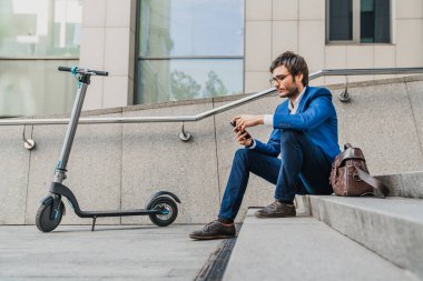 Merdivenlerde elektrikli scooter 'ın yanındaki telefonu kullanan genç işadamının yan görüntüsü