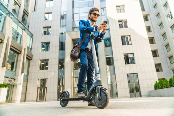 Przystojny Młody Biznesmen Pomocą Smartfona Podczas Jazdy Skuter Elektryczny Pobliżu — Zdjęcie stockowe