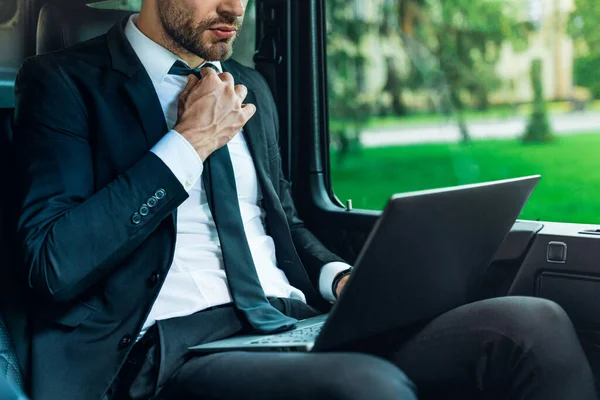 Cropped image of young man in full suit working on laptop and adjusting tie while sitting in the car