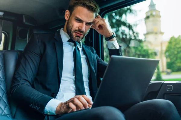 Thoughtful young man in full suit working using laptop and holding his head by hand while sitting in the car