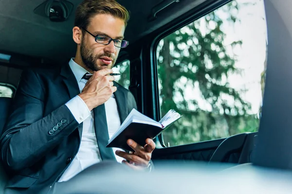 Handsome young man in full suit holding his personal organizer and looking at it while sitting inside of the car