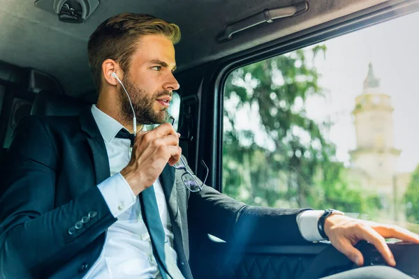 Thoughfull businessman wearing earphones and listening musing while driving in a car with eyeglasses in hand