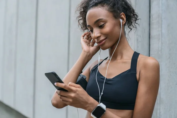 女跑步者一边调整耳机 一边在智能手机上选择音乐 同时停止跑步锻炼 — 图库照片