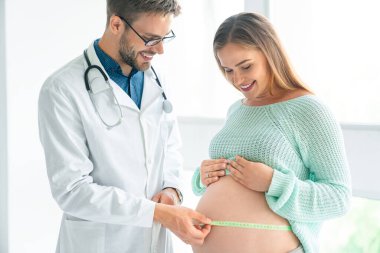 Bir yandan hamile bir kadının karnını teşhir ederken, bir yandan da doktor içeride, hastanede bebeğin büyümesini izlemek için bir mezura kullanıyor..