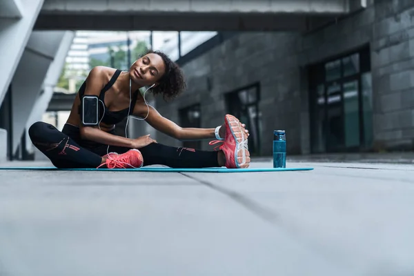 戴耳机的年轻女性在室外健身垫上锻炼和伸展身体 — 图库照片