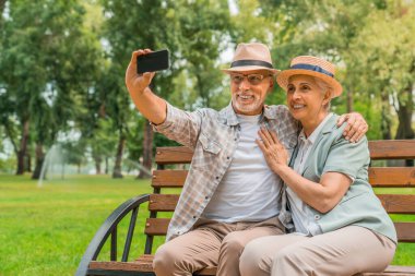 Şapkalı son sınıf çiftimiz dışarıda bankta otururken cep telefonlarıyla selfie çekiyorlar.