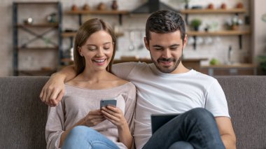 Genç mutlu çift evde kanepede otururken sarılıyor ve akıllı telefon, dijital tablet kullanıyor.