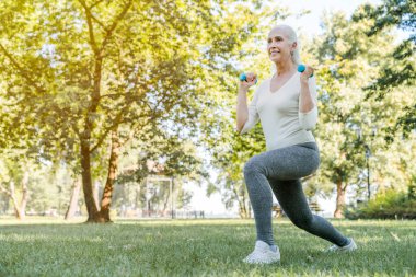 Elinde dambıl olan olgun bir kadın parkta egzersiz yapıyor.