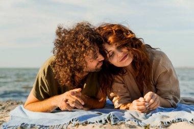 Mutlu gülümseyen beyaz çift kumsalda birbirine aşık ve dinleniyor. Bir adam battaniyeye uzanmış bir kadını öpüyor.