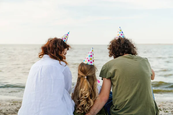 Rear view of caucasian family with party hats having fun on the beach. Man and woman celebrating daughter\'s birthday.